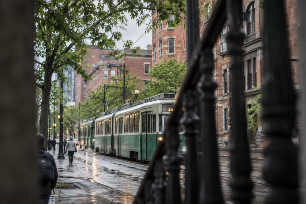 A green line trolley running along a city avenue with brick buildings behind it