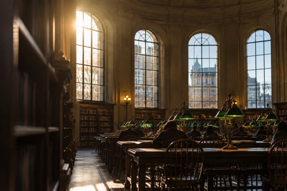 A quiet reading room with tall arched windows and green lamps over long tables
