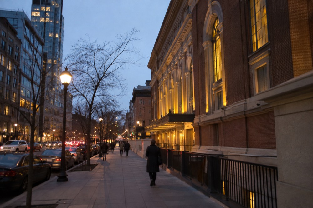 A city avenue at dusk with lit shopfronts and a brick concert hall on the corner