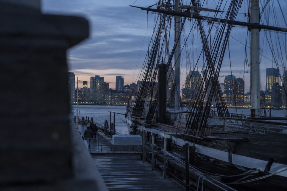 The wooden warship USS Constitution docked at a pier in Charlestown Navy Yard with Boston's skyline behind it