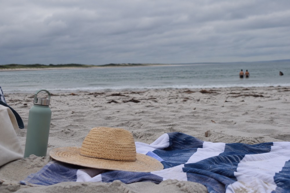A beach towel, sun hat and water bottle set out on sand beside calm water