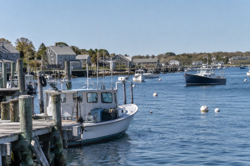 A harbor in a Cape Cod town with fishing boats moored and low buildings along the shore