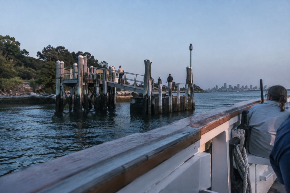 A wooden ferry approaching a New England harbor island dock on a clear summer morning