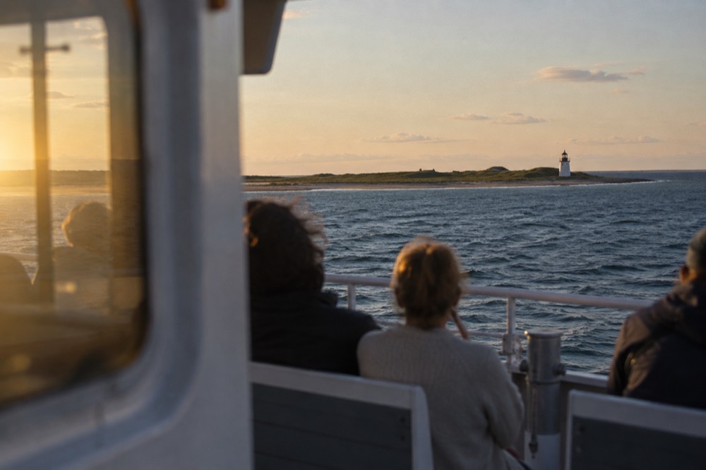 A small ferry crossing open blue water toward a low green island with a lighthouse on the point