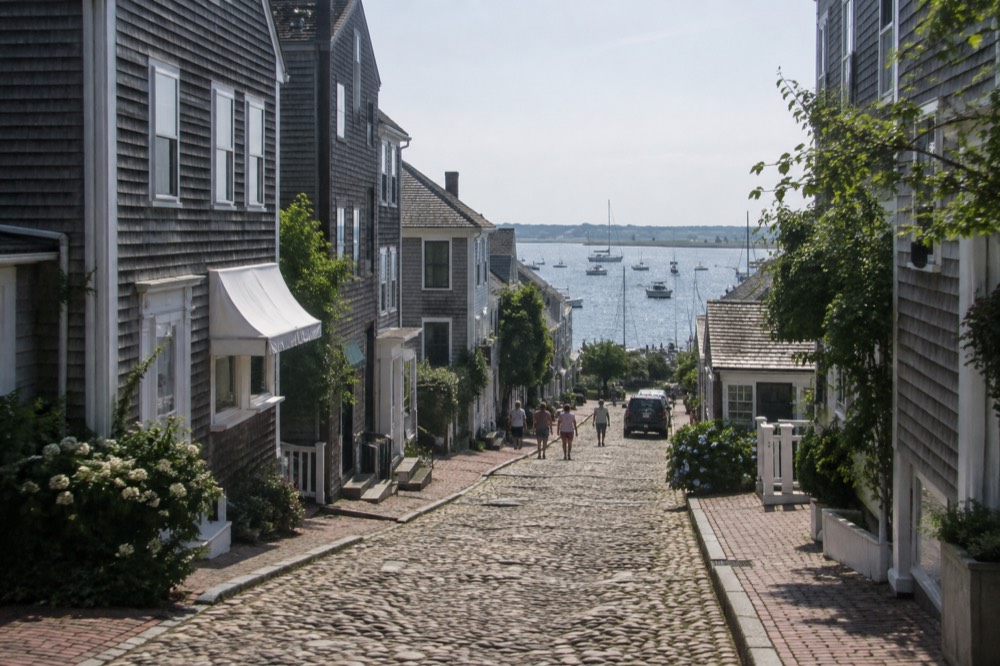 A cobblestone street lined with gray shingled buildings and shop awnings on a sunny afternoon