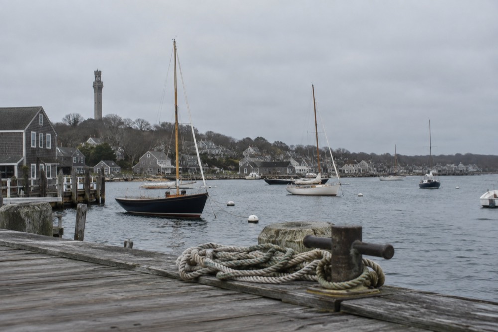 Weathered wooden buildings and a stone tower above a harbor filled with sailboats