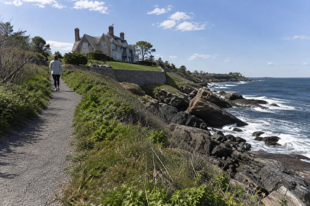 A walker on a coastal path with ocean waves breaking against rocks below a mansion lawn