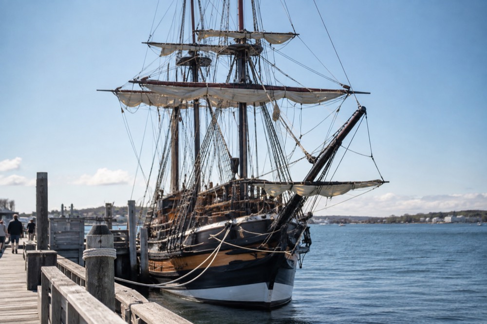 A full-rigged wooden ship with dark hull and furled sails tied at a pier