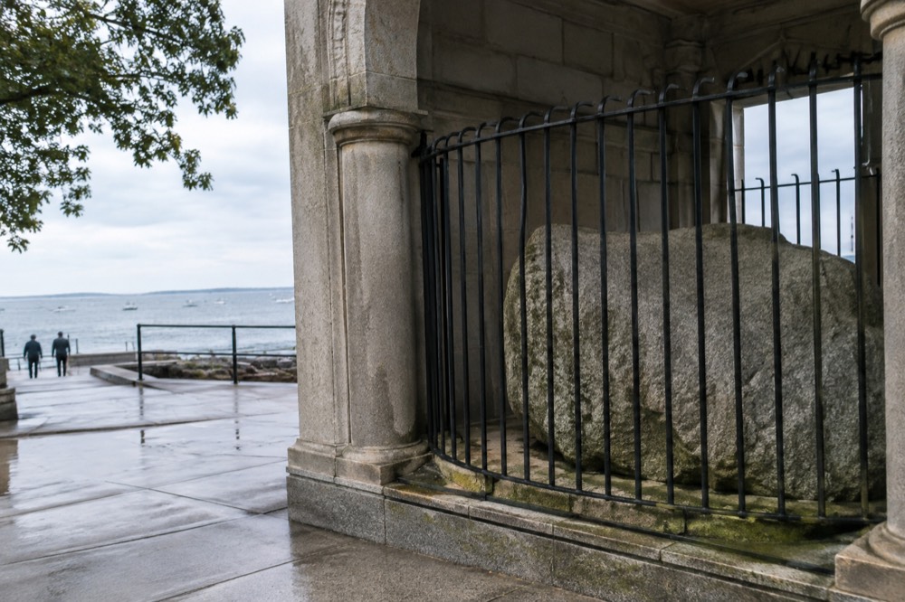A gray glacial boulder displayed behind iron bars in a stone portico at the water's edge