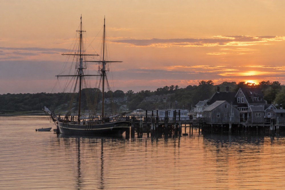 A sailing ship and harbor buildings at dusk seen across calm water