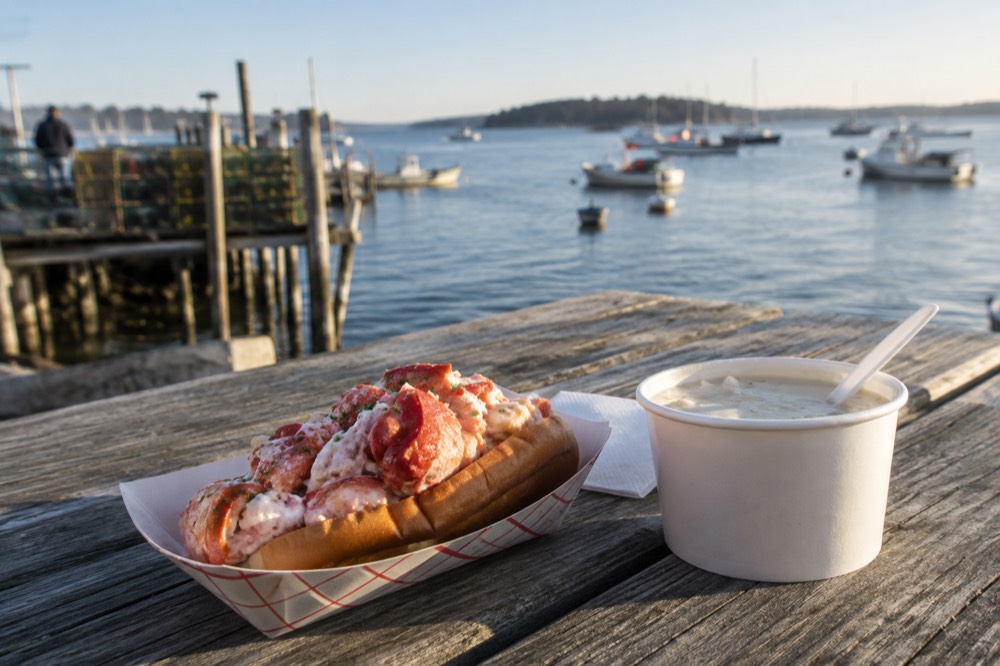 A lobster roll on a paper tray beside a paper cup of chowder