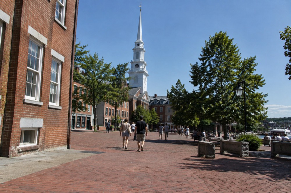 A town square with brick buildings, a church spire and people walking