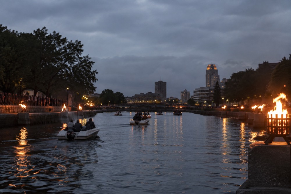 Small boats on a city river at night with lit fires along the banks