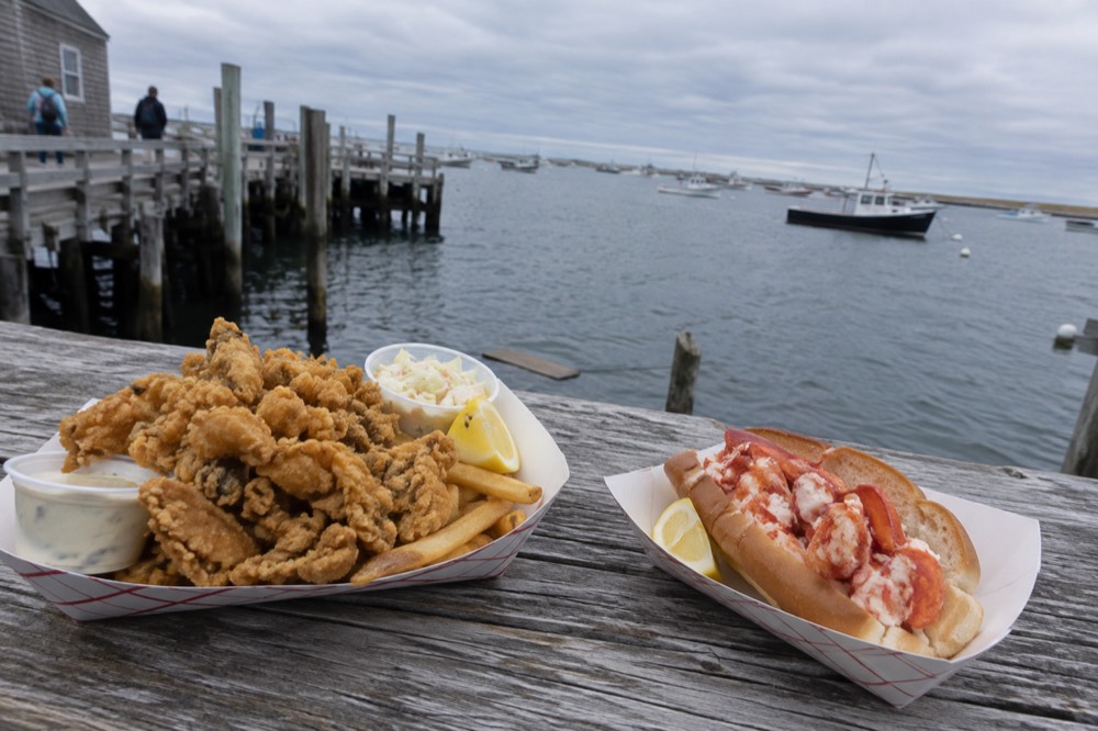 A plate of fried clams and a lobster roll on a picnic table by the harbor