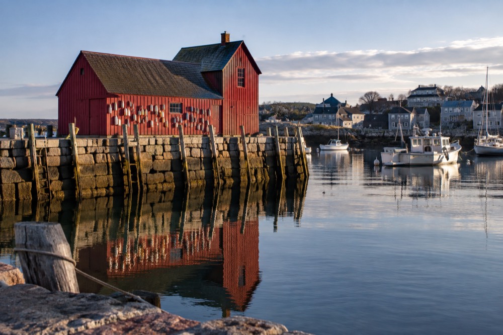 A red wooden fishing shed on a stone wharf reflected in calm harbor water