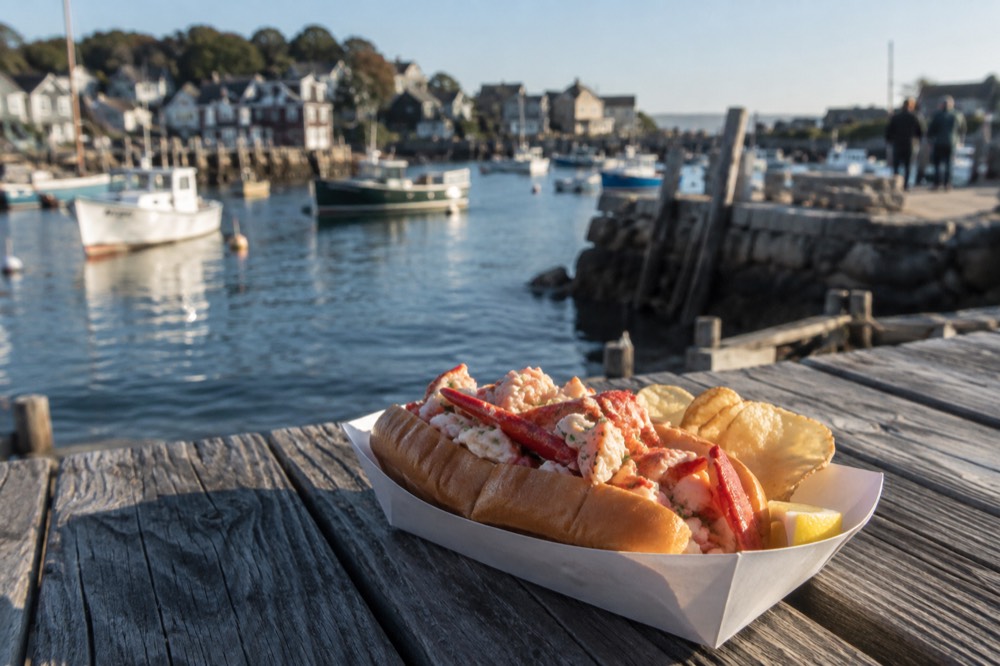 A lobster roll served on a wooden picnic table beside a harbor