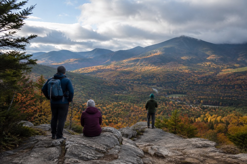Hikers in warm jackets standing at a scenic overlook above a valley of autumn trees