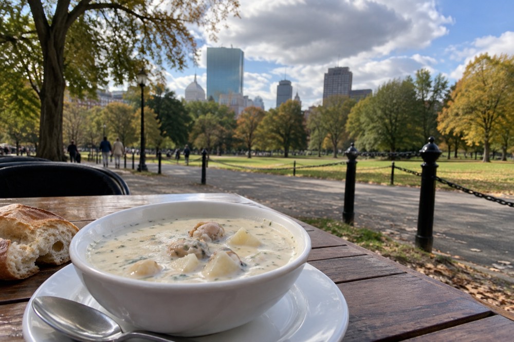 A white bowl of creamy New England clam chowder with clams and potatoes on a wooden restaurant table