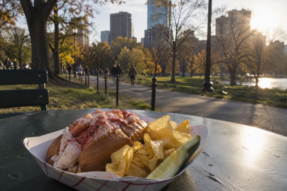 A lobster roll served with chips and a pickle on a paper tray at an outdoor table