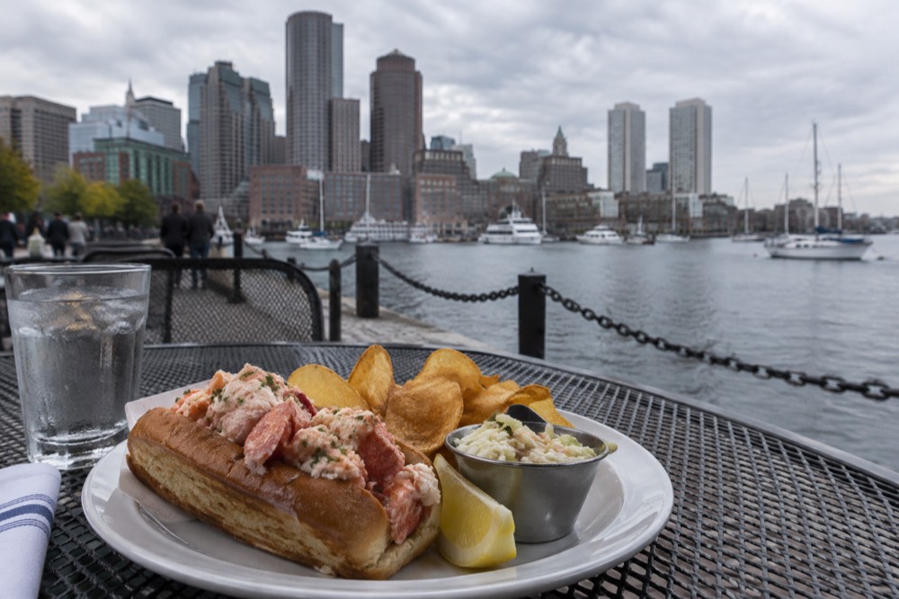 A patio table with a lobster roll plate beside a harbor view in afternoon light