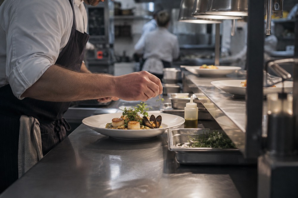 A chef plating a seafood dish at a pass in a Boston restaurant kitchen