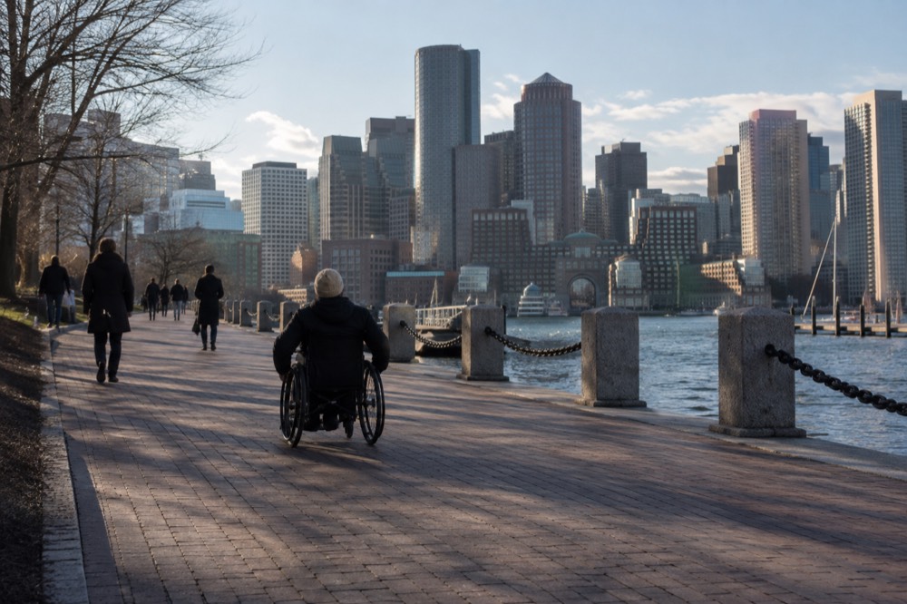 A wheelchair user traveling along a wide brick sidewalk near Boston Harbor with the city skyline behind