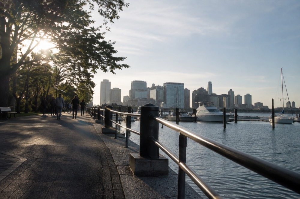 A paved harborfront walkway with railing, water, and boats at a wharf