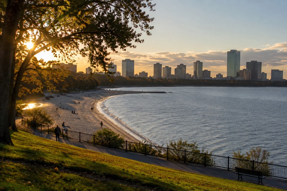 A wide sandy beach with calm water, a curved shoreline and a city skyline in the distance