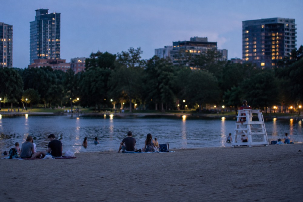 Families sitting on towels on a sandy urban beach with a lifeguard chair and calm water