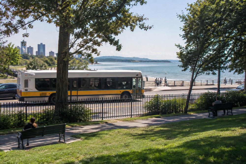 A public transit bus stopped at a beachfront street with sand and water beyond a rail fence