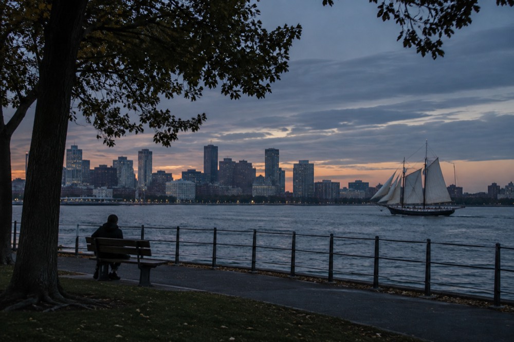 A wooden tall ship under sail on open harbor water with a city skyline behind it