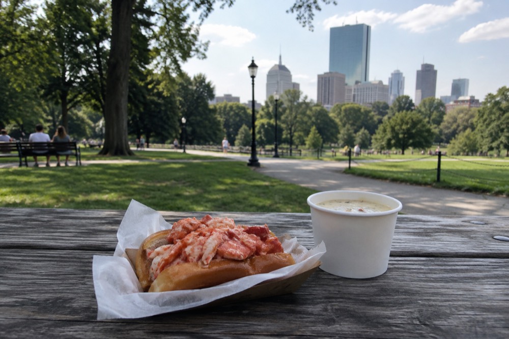 A lobster roll sandwich on a wooden picnic table beside a paper cup of chowder