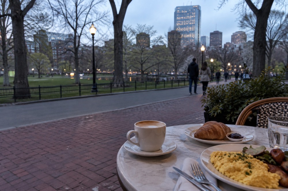 A sunny café table with coffee, pastries and eggs, set beside a brick sidewalk in an old Boston neighborhood