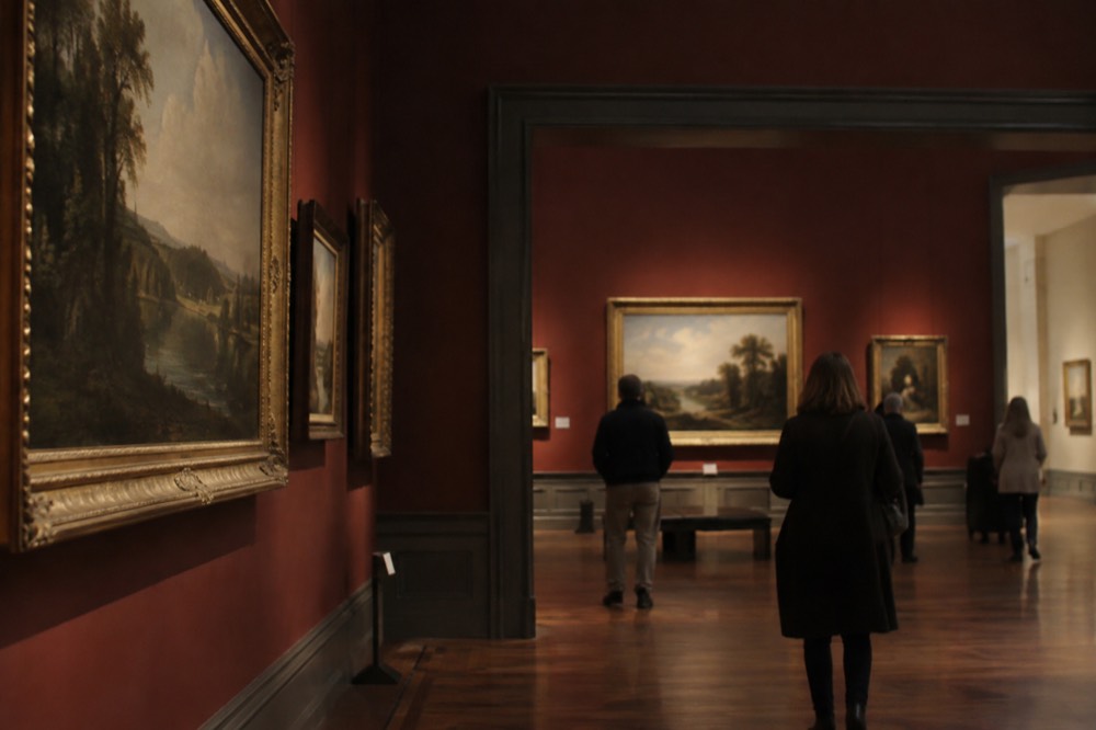 A museum gallery with paintings hung on deep red walls and visitors walking through