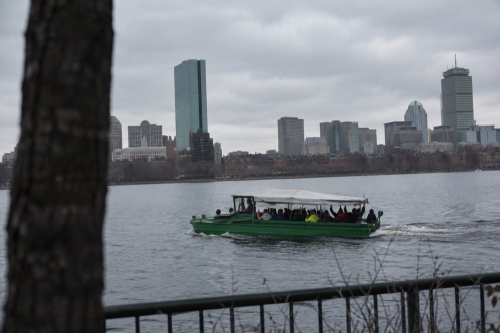 Passengers waving from a duck boat floating on the Charles River with the Back Bay skyline behind