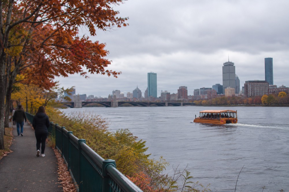 A duck boat on the Charles River in fall with orange foliage along the riverbank