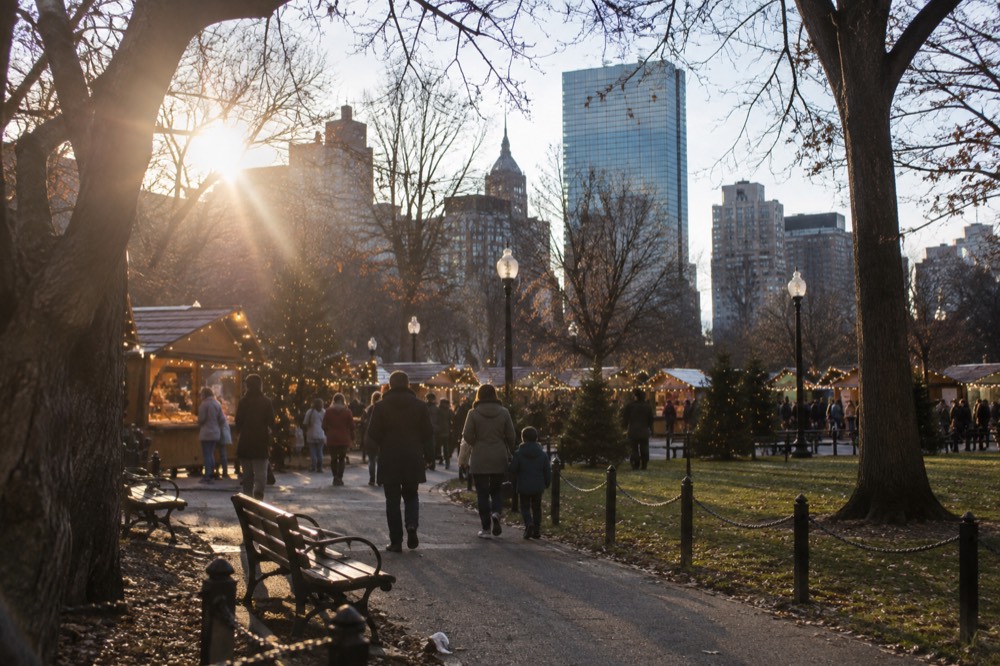 Families strolling a holiday market with string lights between brick buildings