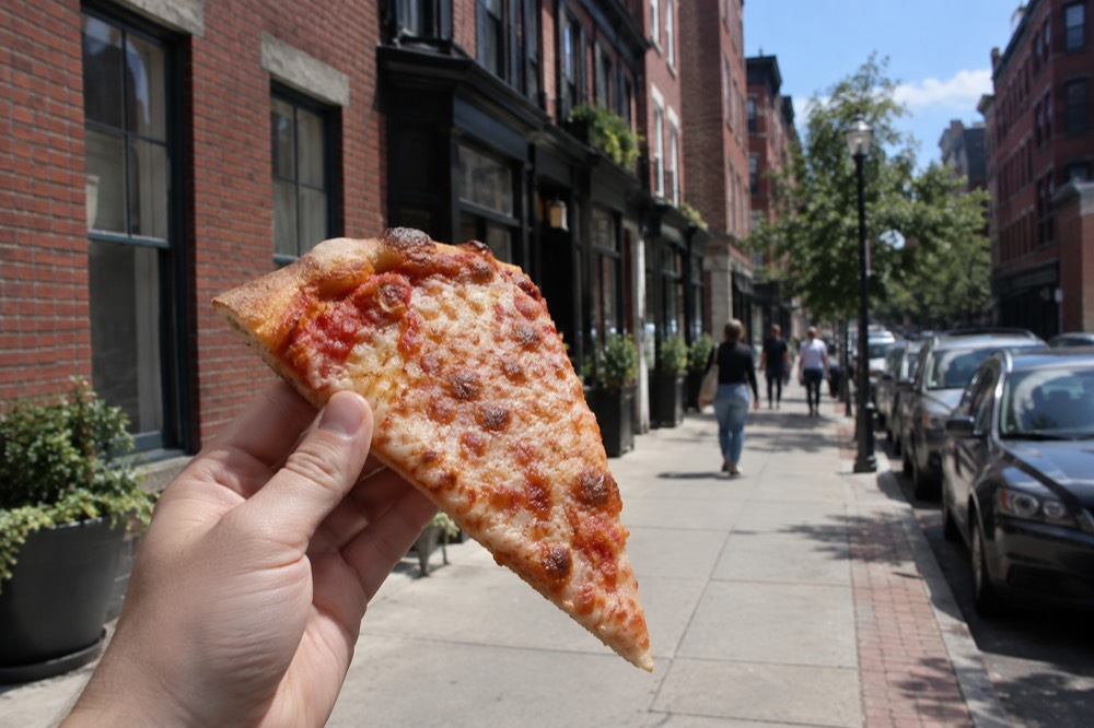 A slice of cheese pizza held up in front of a row of North End brick storefronts