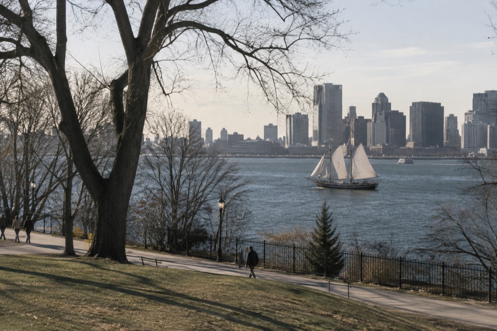 A wooden tall ship with white sails crossing a harbor with a city skyline behind it