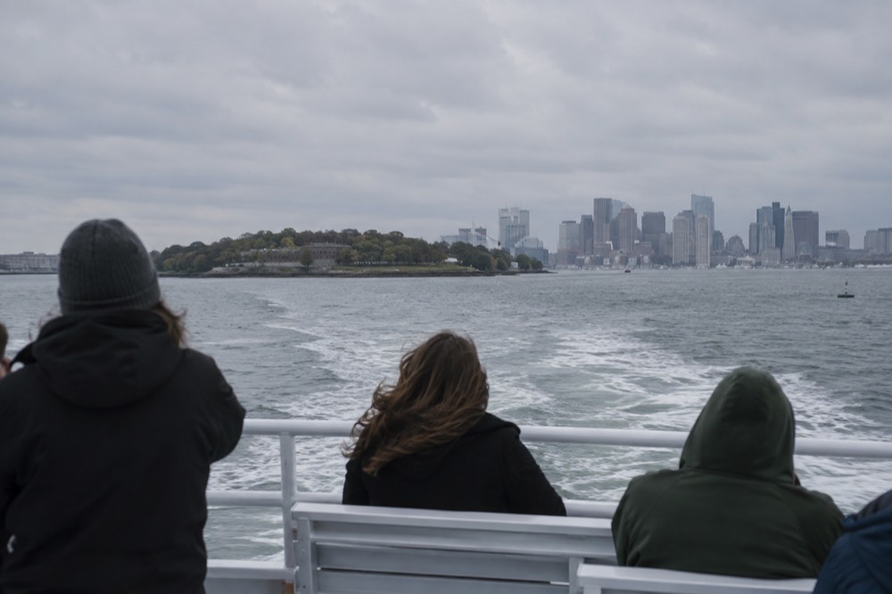 A ferry crossing open harbor water toward a green island with a city skyline behind