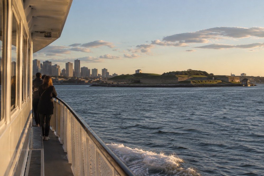 A ferry crossing open harbor water toward a green island with an old fort
