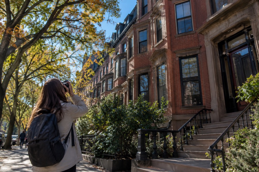 A traveler in a light jacket photographing brownstone row houses on a tree-lined Boston street