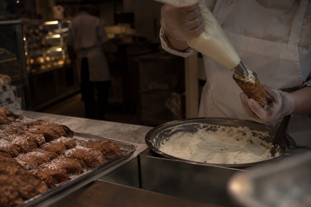 A cannoli being filled behind a bakery counter at night