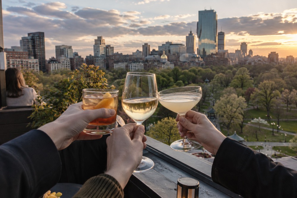 Friends toasting with drinks on a rooftop overlooking the lit Boston skyline