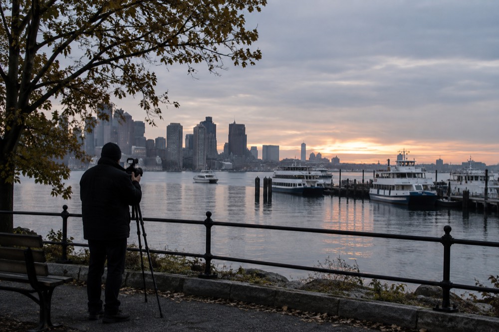 Photographer with a camera on a tripod overlooking the Boston harbor at dawn with ferries at their docks