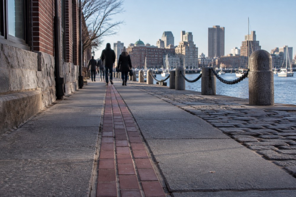 A red brick Freedom Trail line running along a sidewalk toward Boston harbor buildings