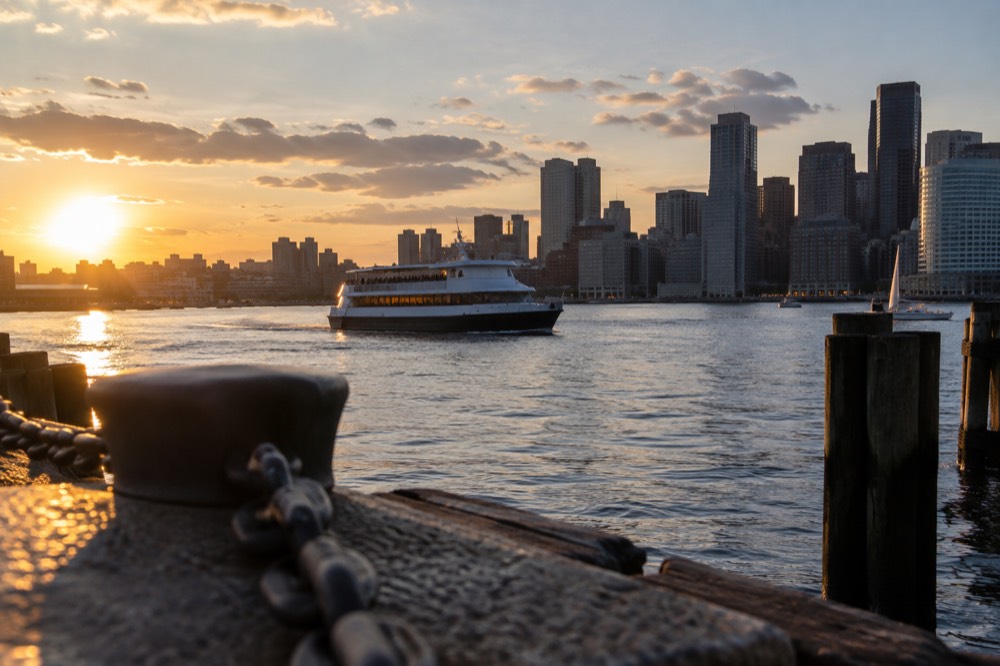 A Boston harbor waterfront at sunset with a cruise boat passing the city skyline