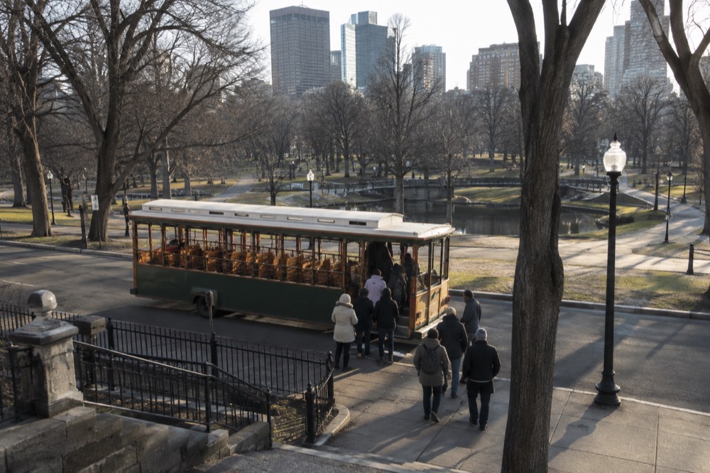 Passengers boarding an open-air trolley at a curb stop near a Boston plaza