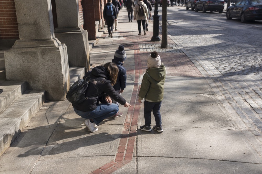 A parent pointing out a red-brick Freedom Trail line embedded in a Boston sidewalk to two children