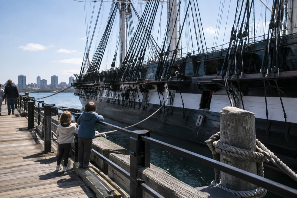 Children looking up at the masts of an old wooden warship at a Charlestown pier